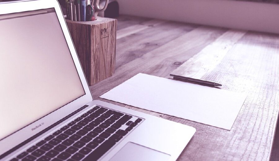 Close up image of an open laptop computer on a desk along with a piece of paper and a pen and a wooden container holding more pens and pencils